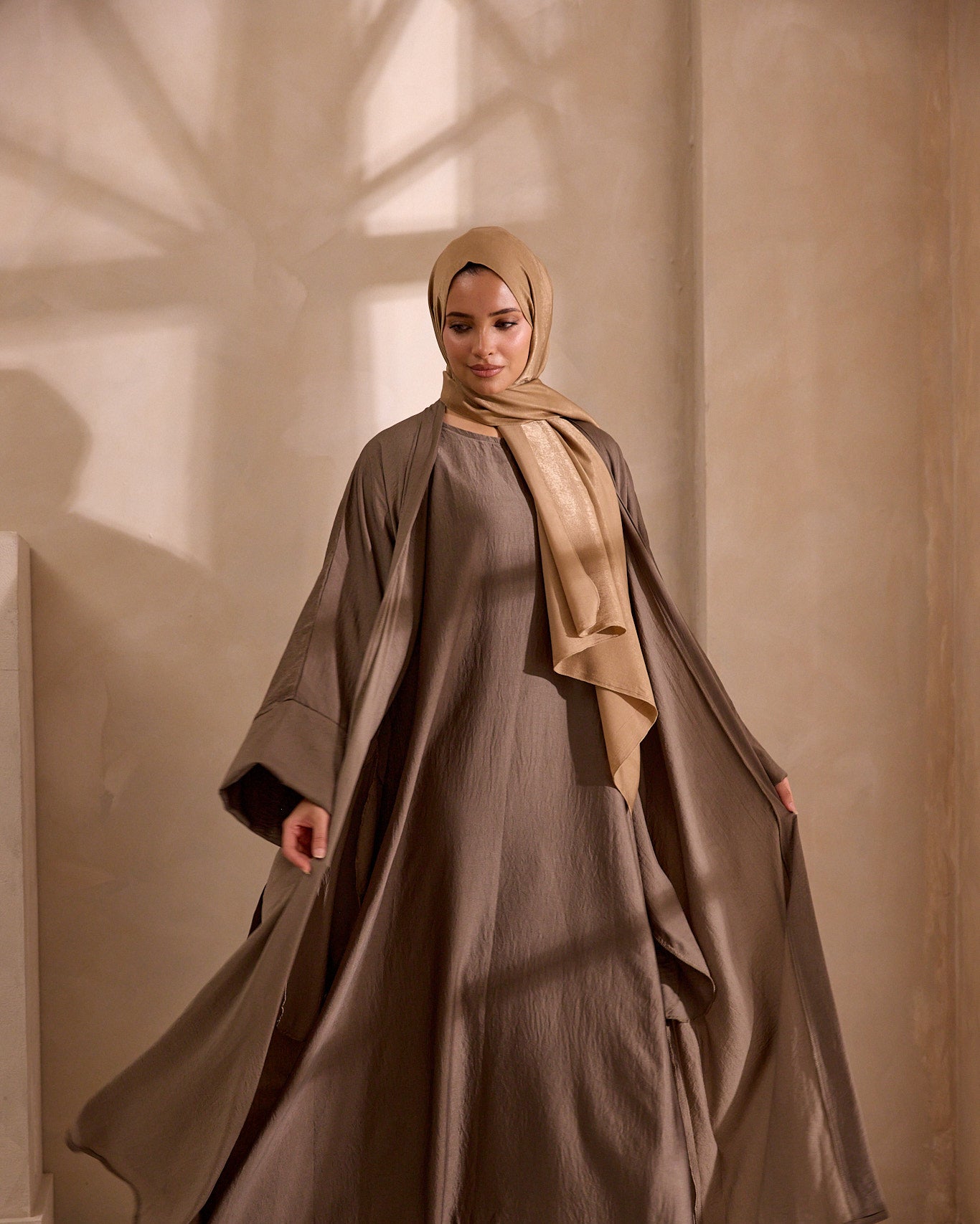 Woman wearing a long, flowing dress with a beige hijab in a softly lit room.