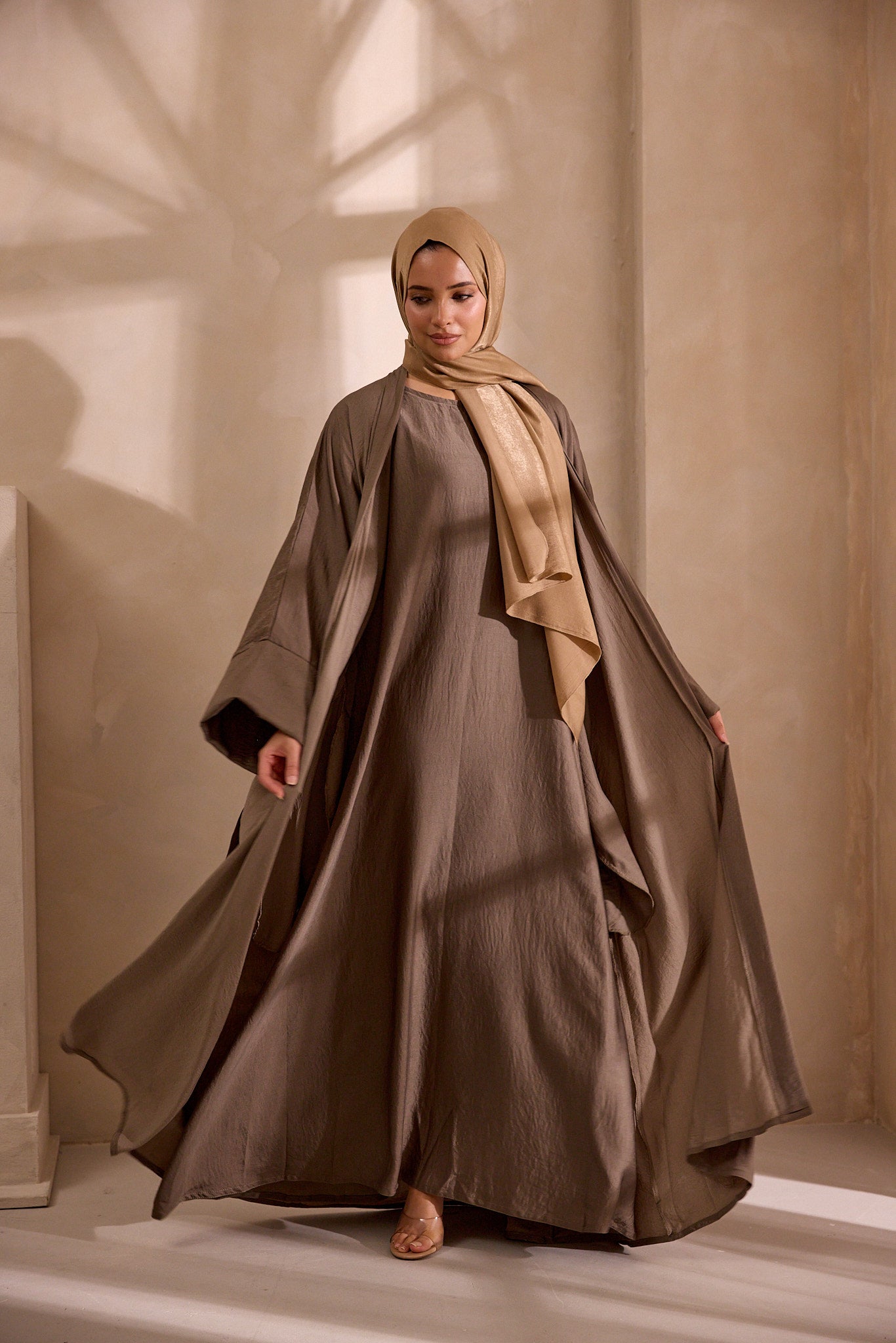 Woman wearing a long, flowing dress with a beige hijab in a softly lit room.