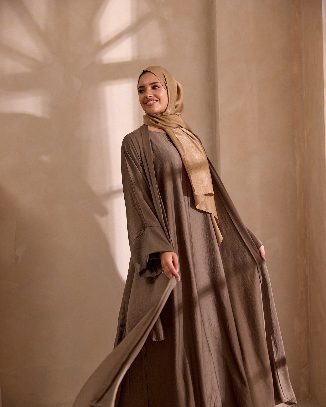 Woman wearing a long brown dress and beige hijab standing against a neutral background