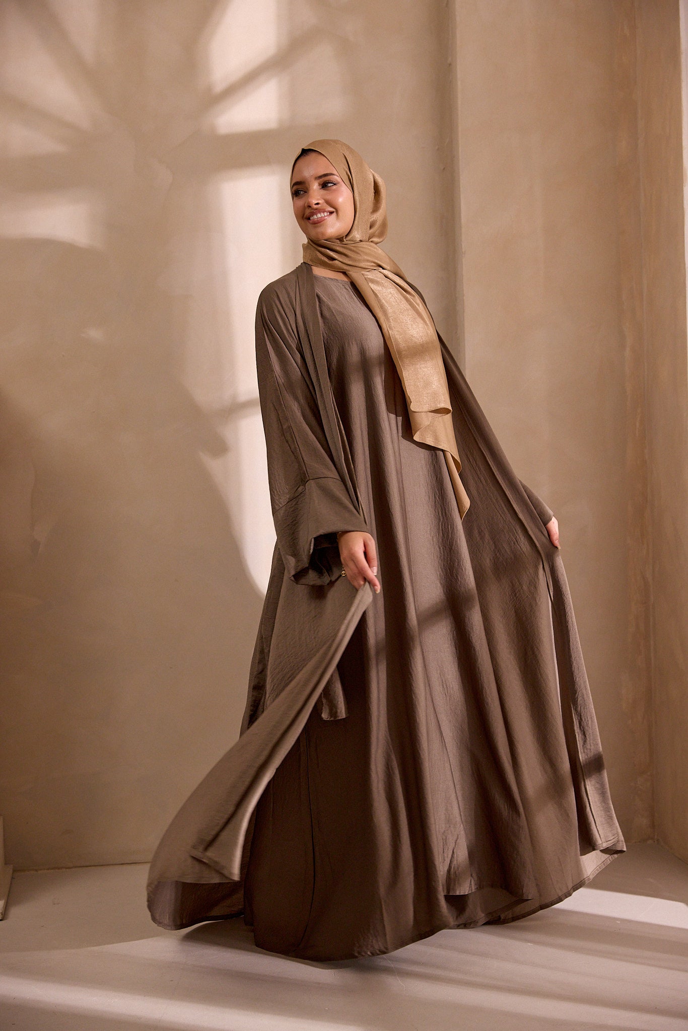 Woman wearing a long brown dress and beige hijab standing against a neutral background