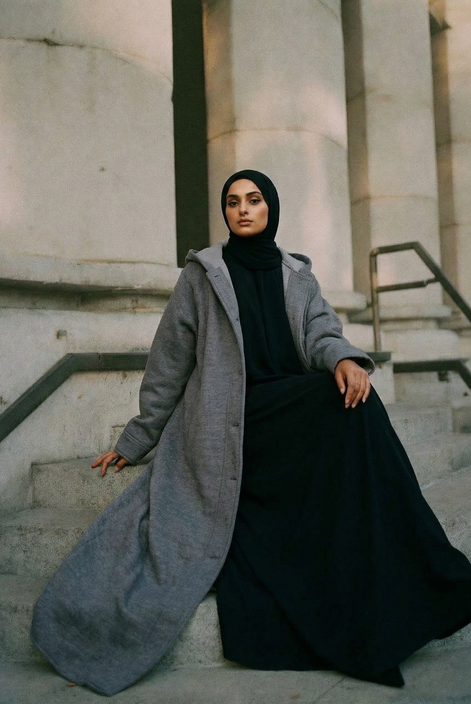 Woman in a long gray coat and black hijab sitting on stone steps.