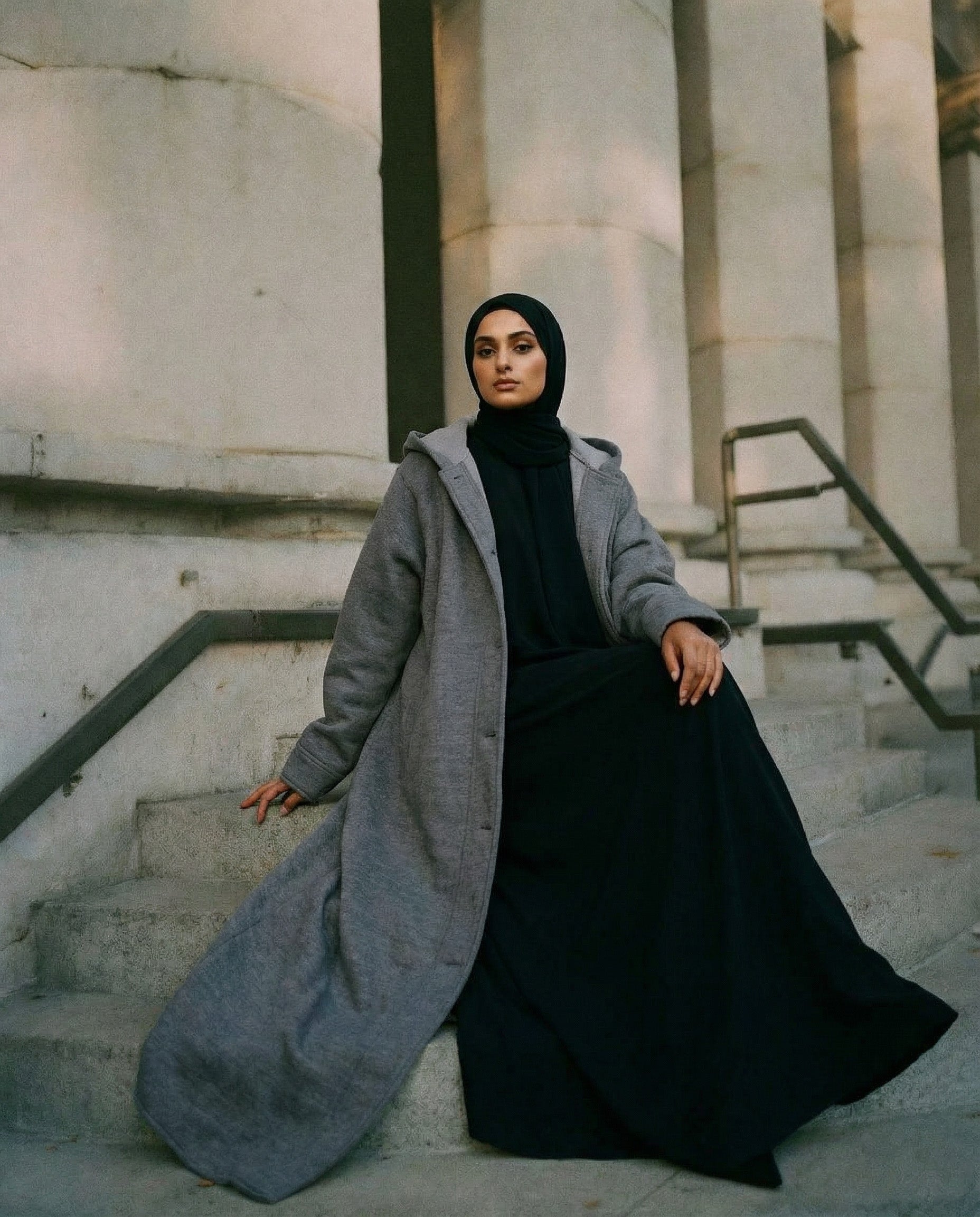 Woman in a long gray coat and black hijab sitting on stone steps.