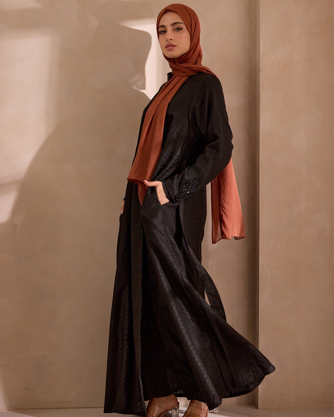 Woman wearing a black outfit with a rust-colored hijab against a beige wall.