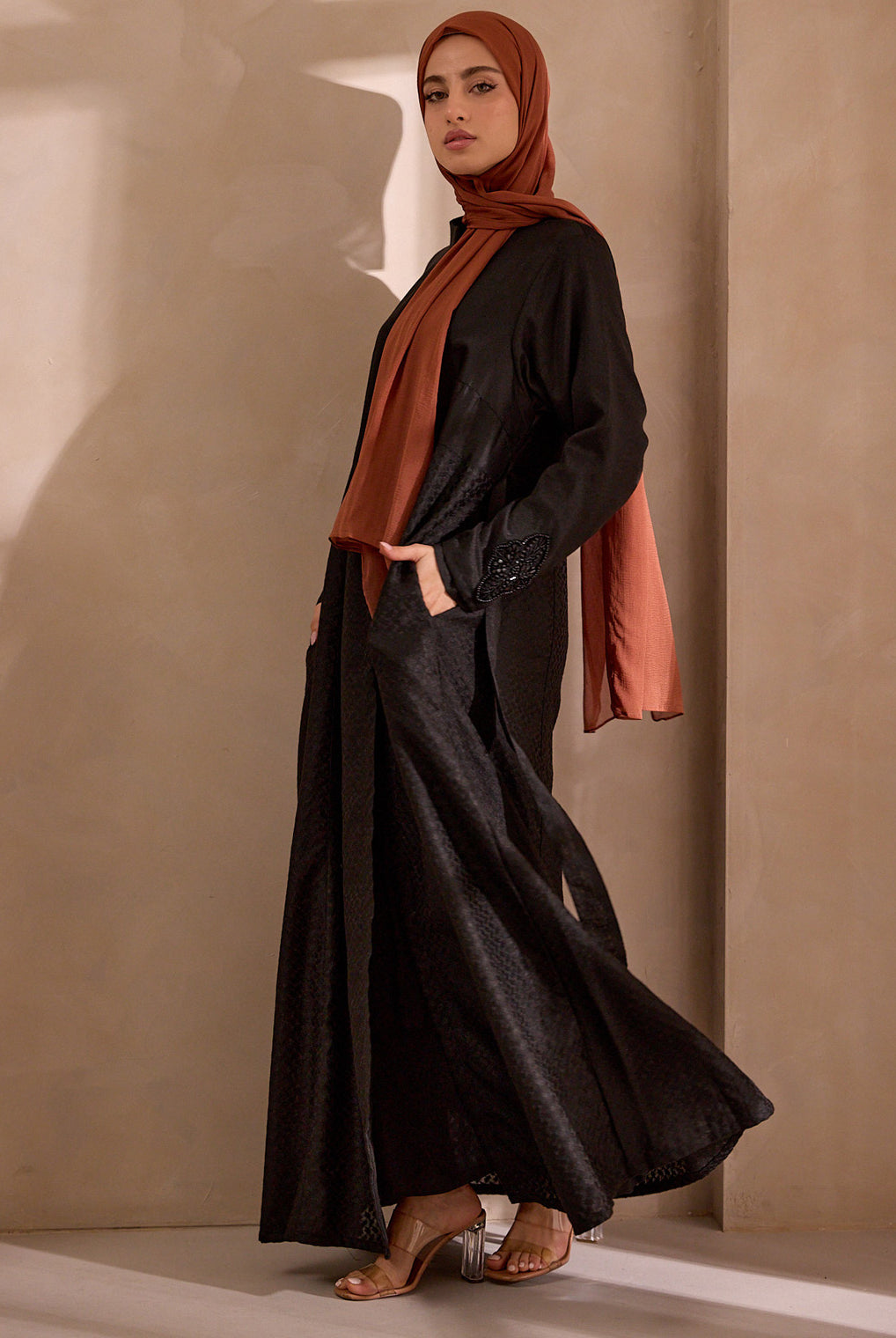 Woman wearing a black outfit with a rust-colored hijab against a beige wall.