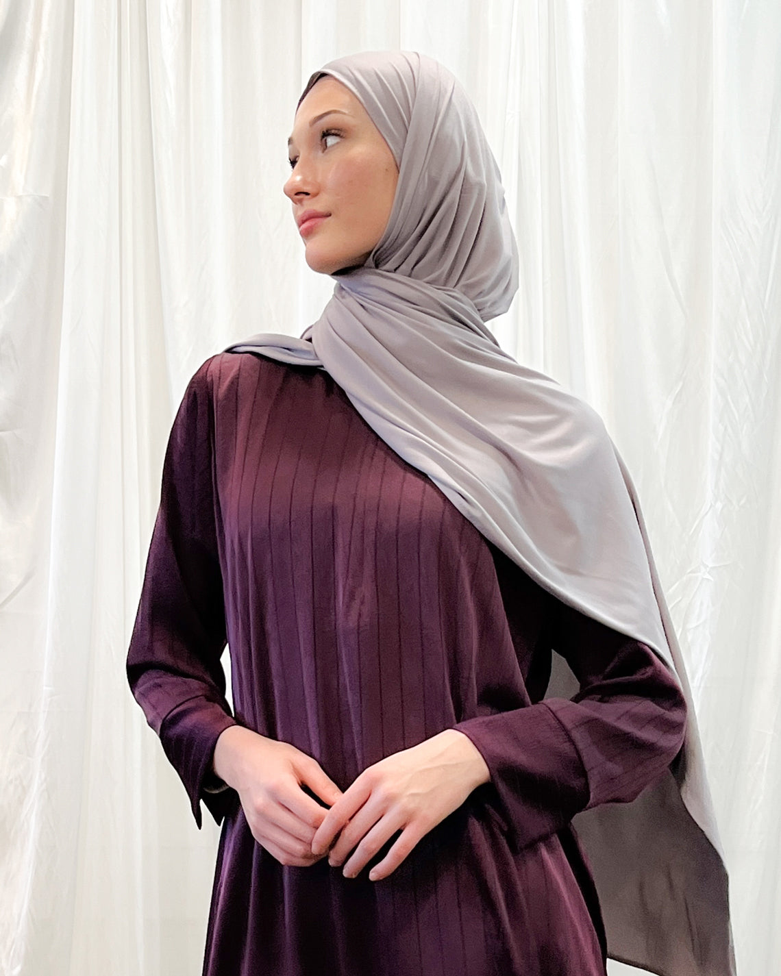 A woman wearing a purple dress and a gray headscarf, standing in front of a white curtain.