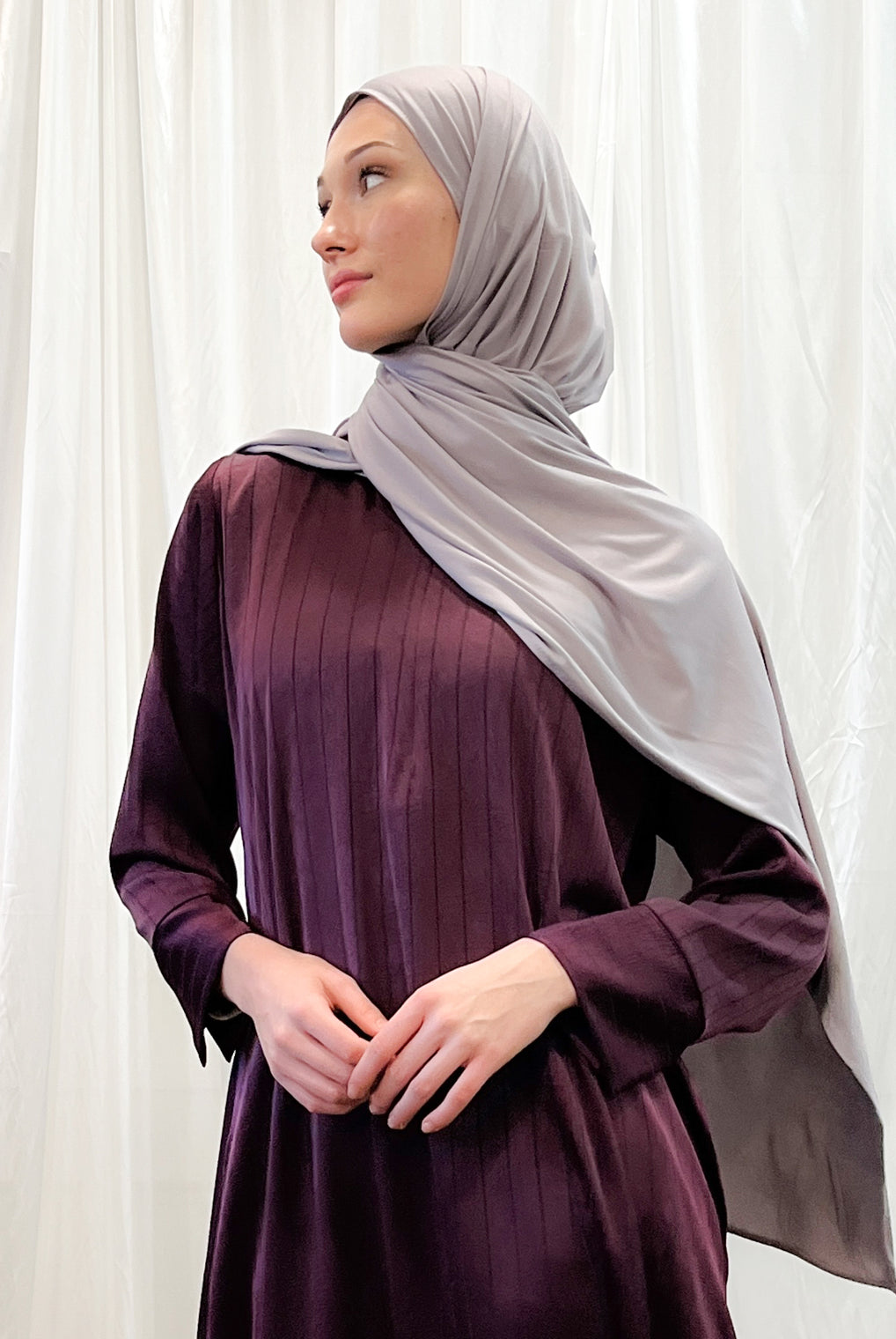 A woman wearing a purple dress and a gray headscarf, standing in front of a white curtain.