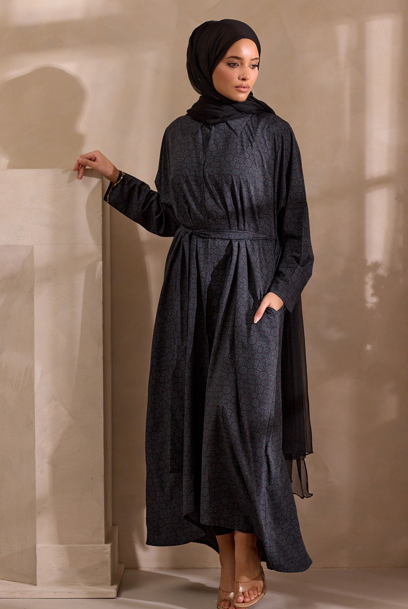 Woman wearing a dark gray dress and hijab against a beige wall.