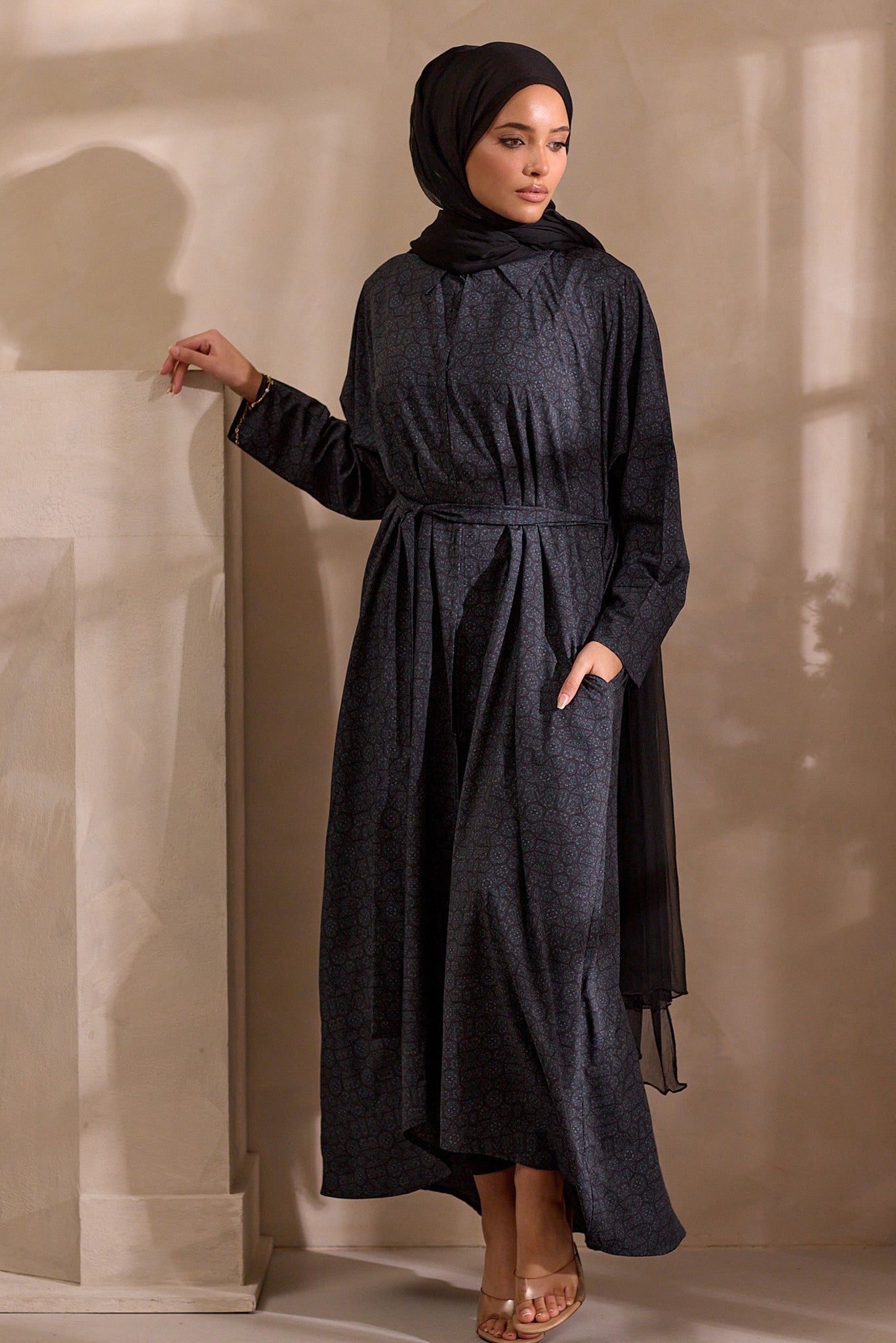 Woman wearing a dark gray dress and hijab against a beige wall.