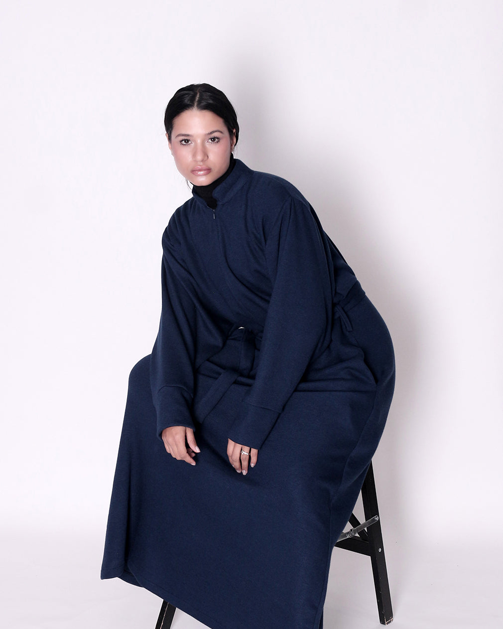 Person wearing a long navy blue garment sitting on a chair against a white background