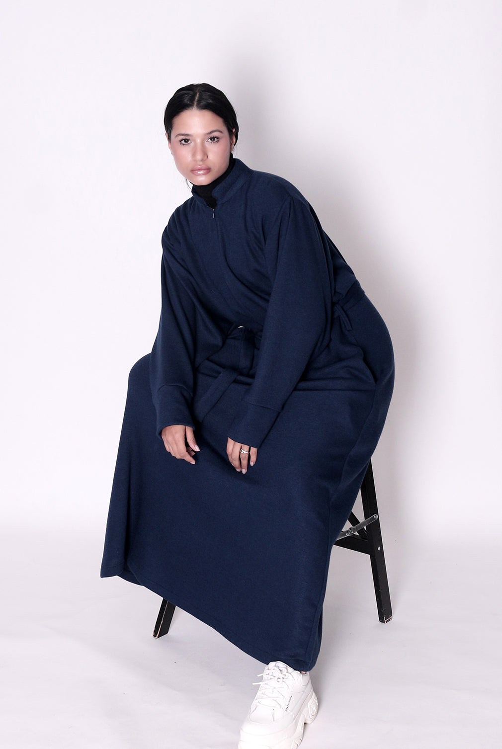 Person wearing a long navy blue garment sitting on a chair against a white background