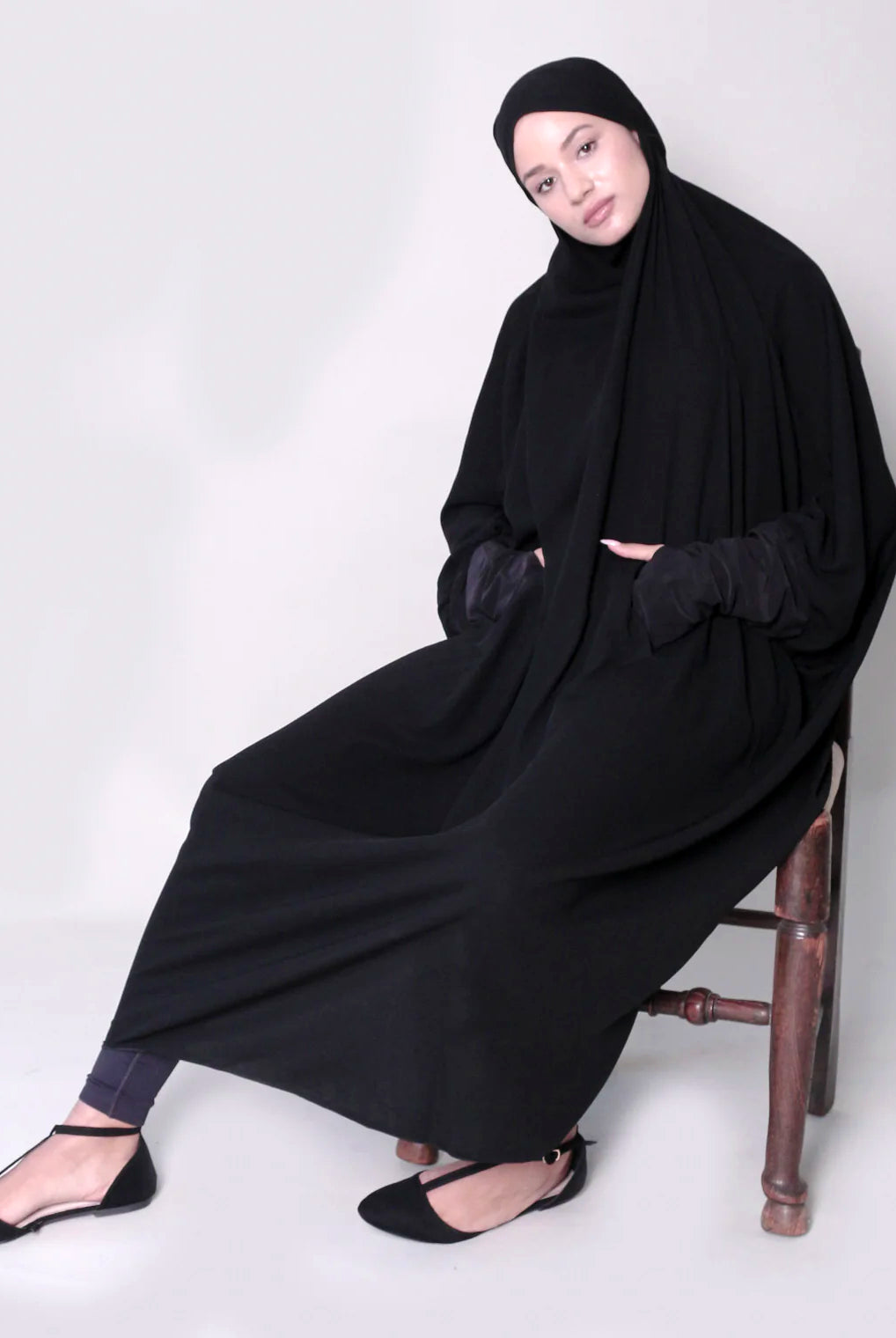 A woman sitting on a chair wearing a black jilbab with thumb holes and a zip at the front.