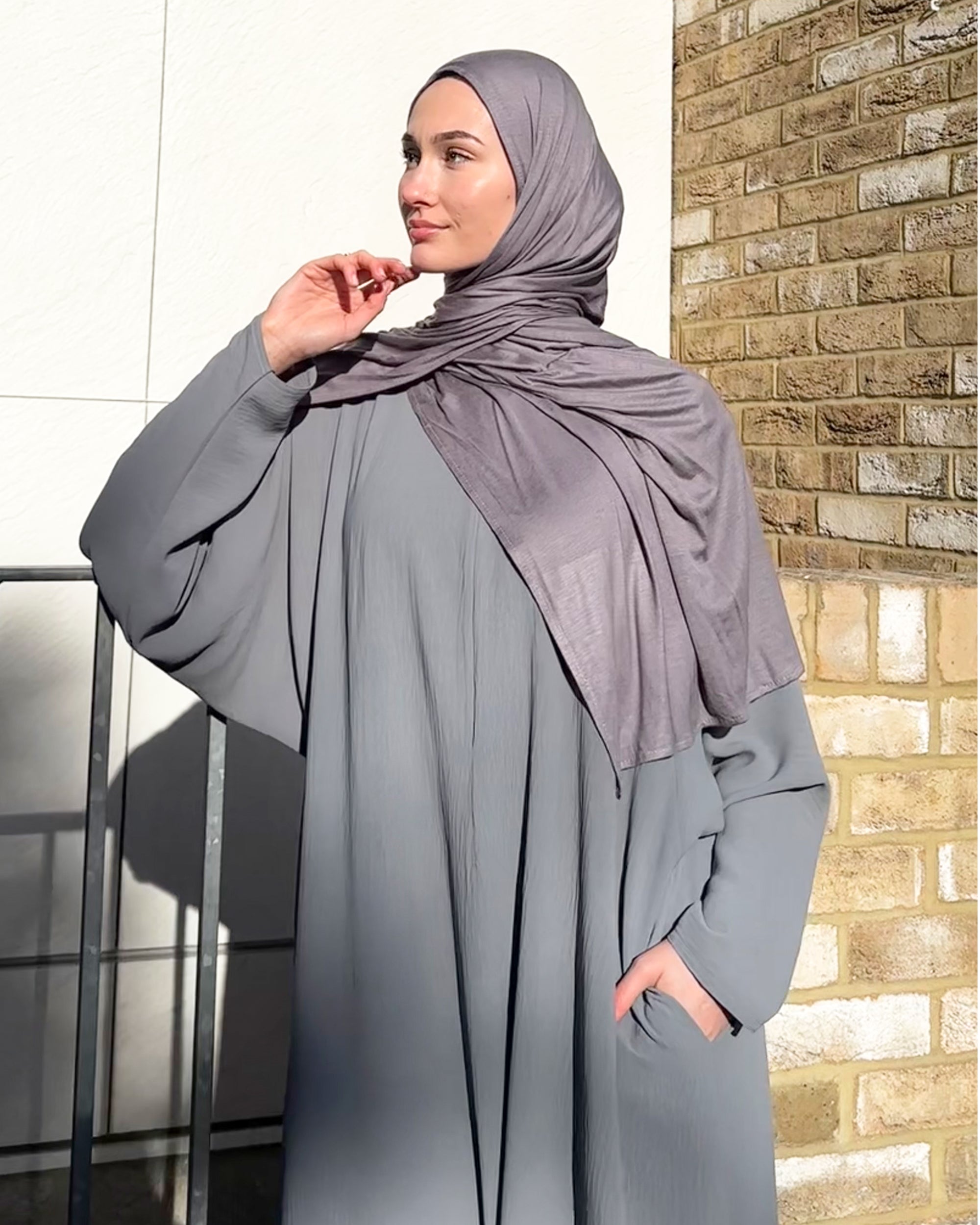 Woman wearing a gray abaya and maxi jersey hijab standing against a brick wall.