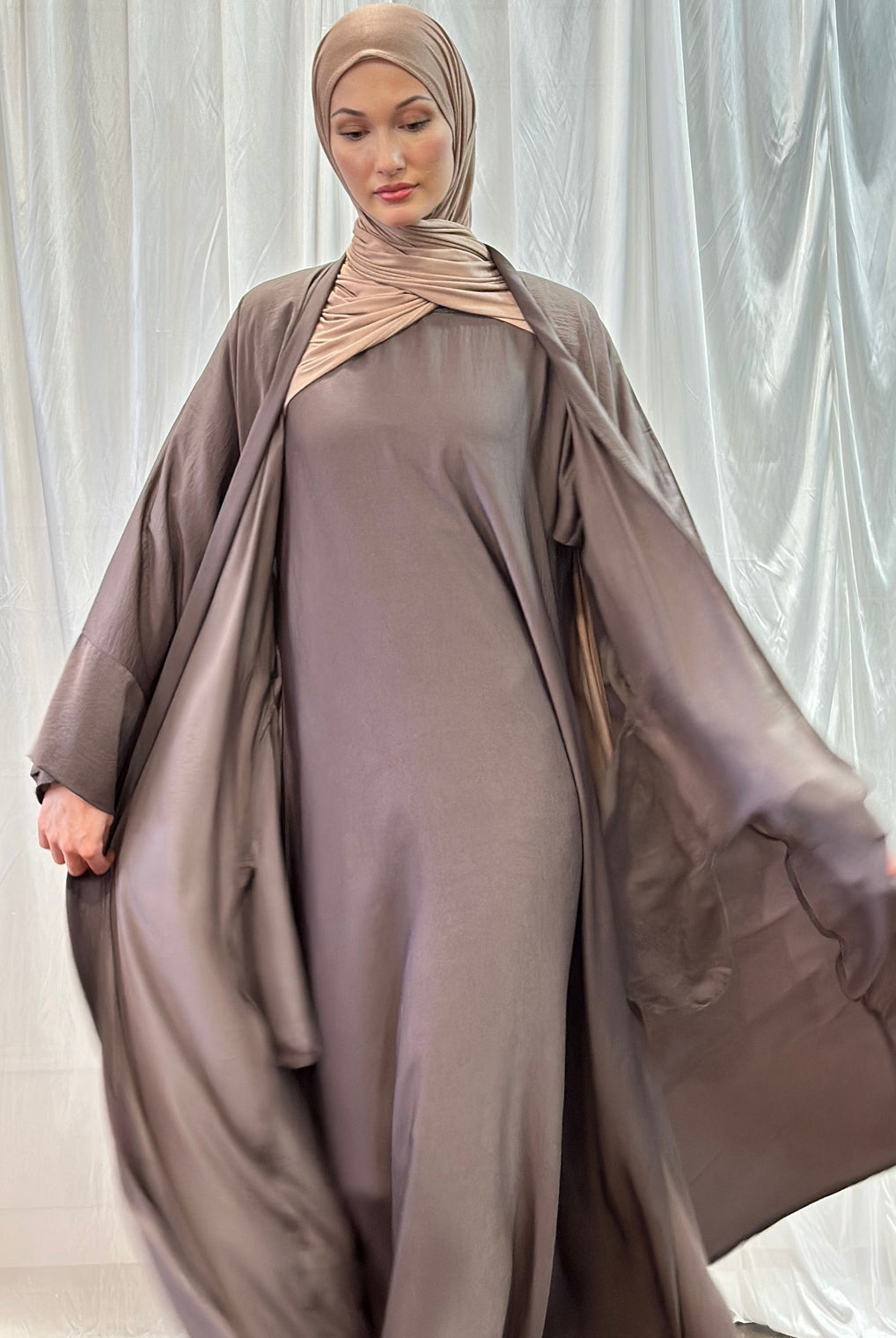 Image depicting a woman wearing a luxurious Hareer Abaya in a neutral brown tone, perfectly complemented by the matching Hareer Jacket. The smooth texture of the silk fabric enhances the graceful drape of the garment, ideal for Spring/Summer fashion.