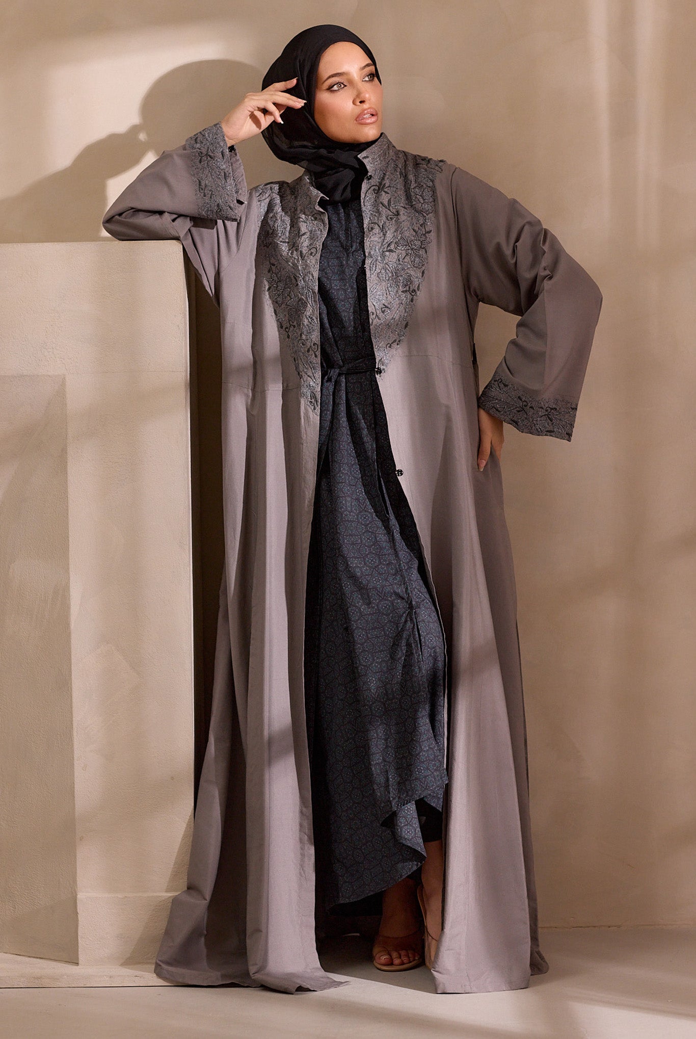 Woman wearing a long, light gray dress with a black hijab against a neutral background open abaya
