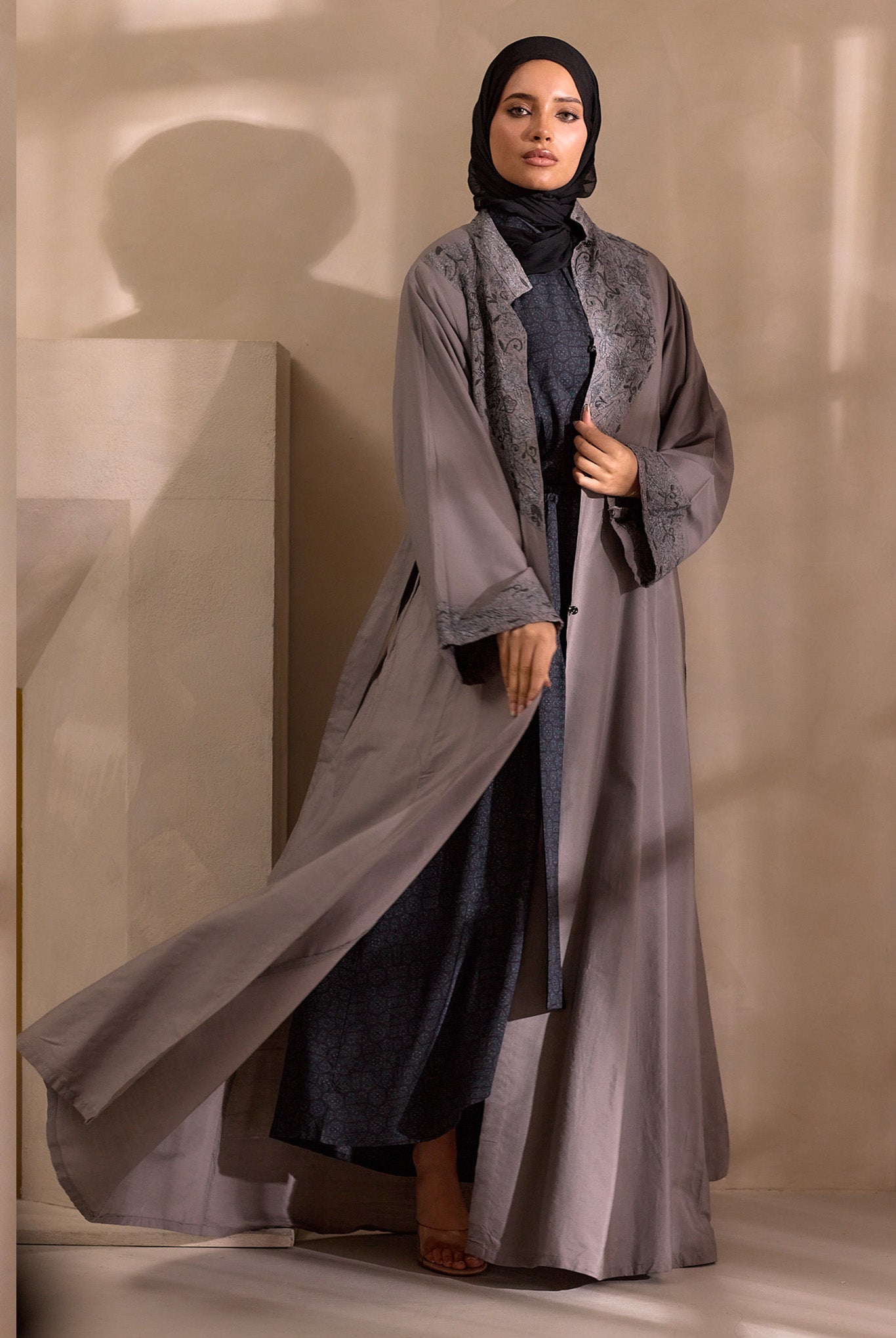 Woman wearing a long, light gray abaya over a dark outfit against a neutral background
