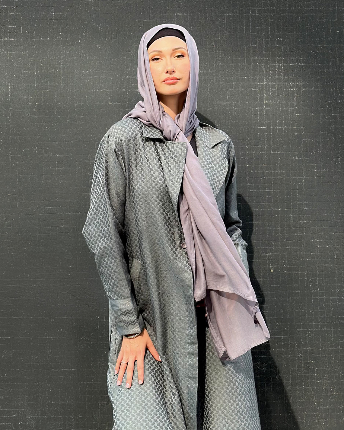 Person wearing a long, light gray khafiyah textured jacket with a matching maxi jersey hijabagainst a dark background