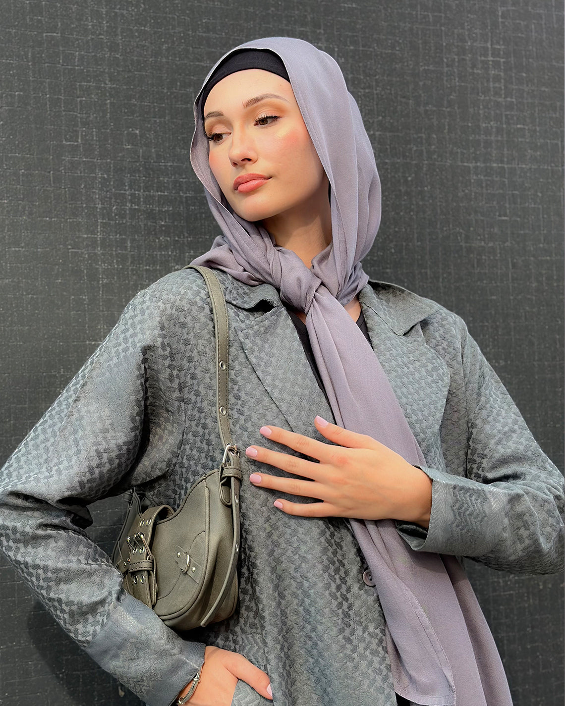 Woman wearing a gray coat and light purple maxi jersey hijab with a green bag against a dark background