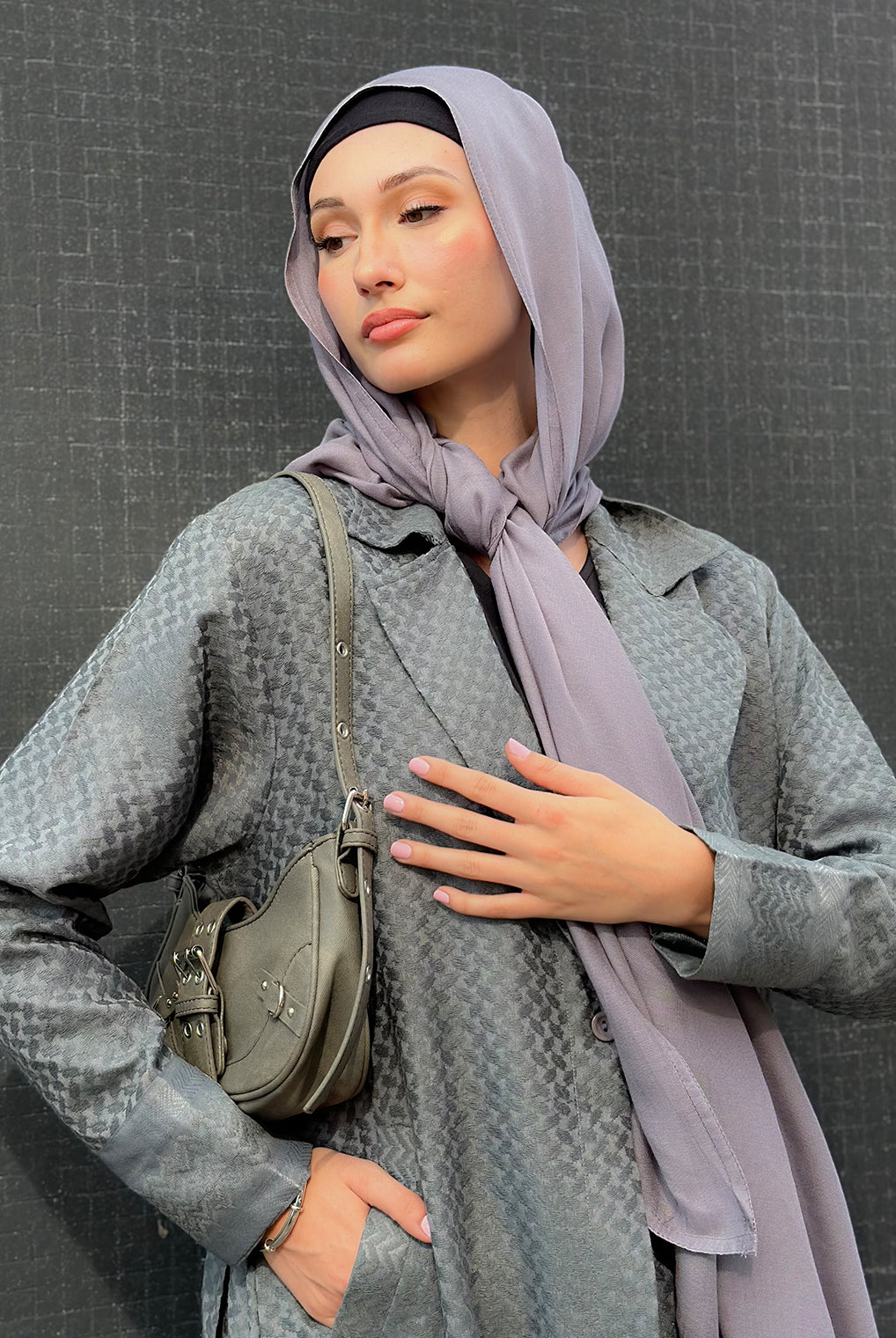 Woman wearing a gray coat and light purple maxi jersey hijab with a green bag against a dark background