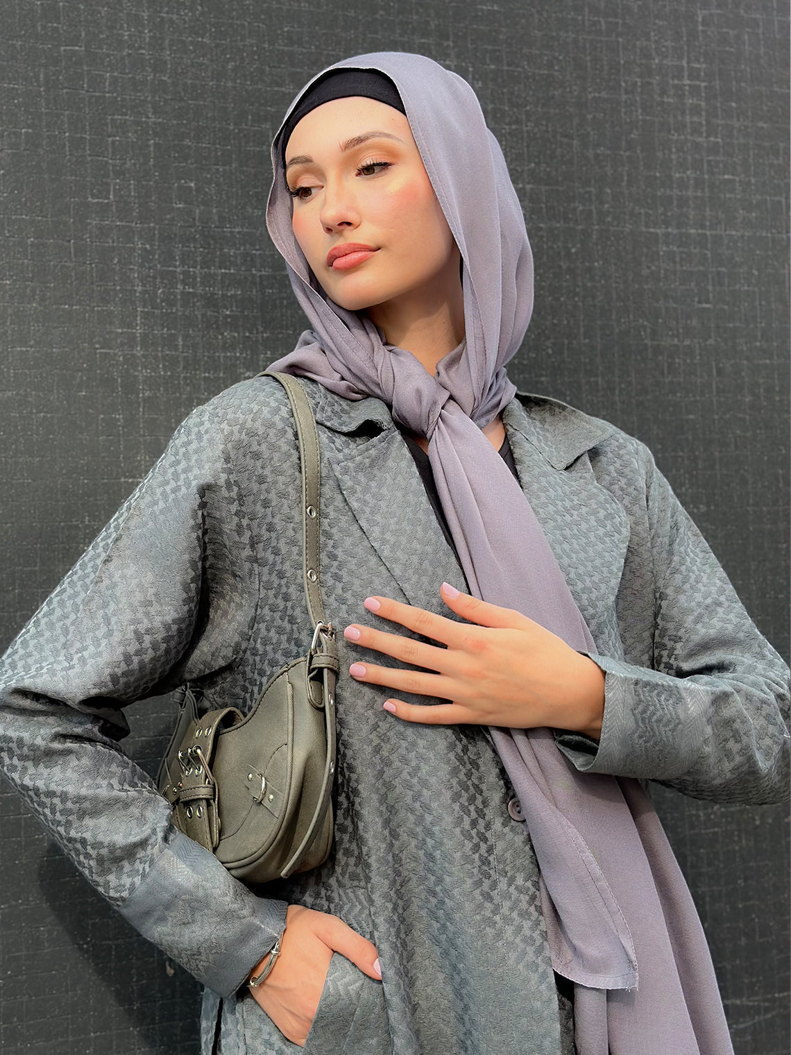 Woman wearing a gray coat and light purple maxi jersey hijab with a green bag against a dark background