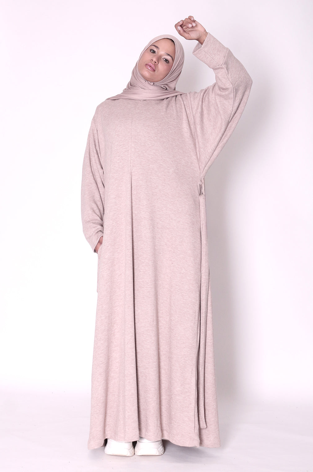 Woman wearing a long, light pink dress knitted abayawith a hood on a white background