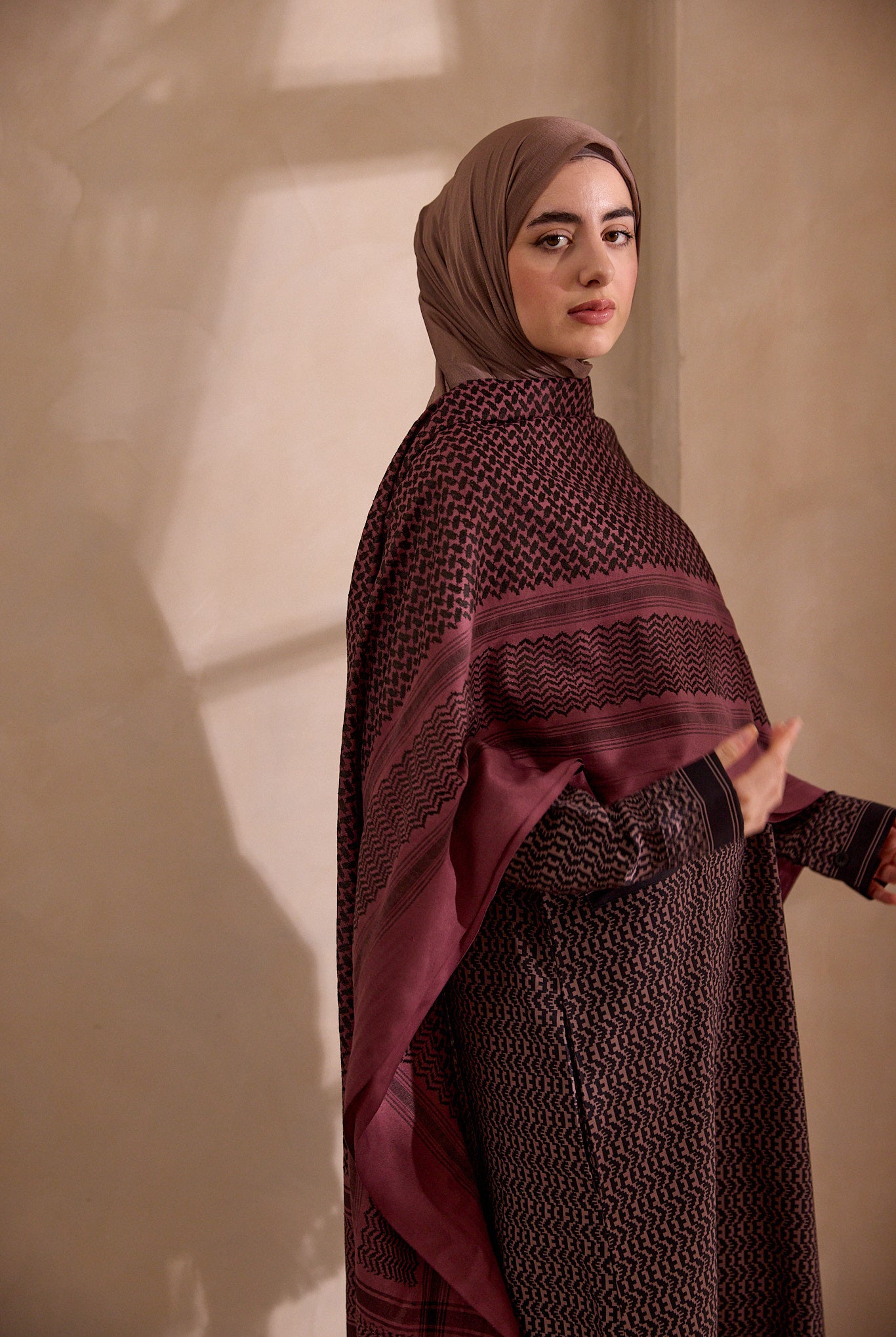 Woman wearing a dark brown hijab and matching outfit against a beige wall.