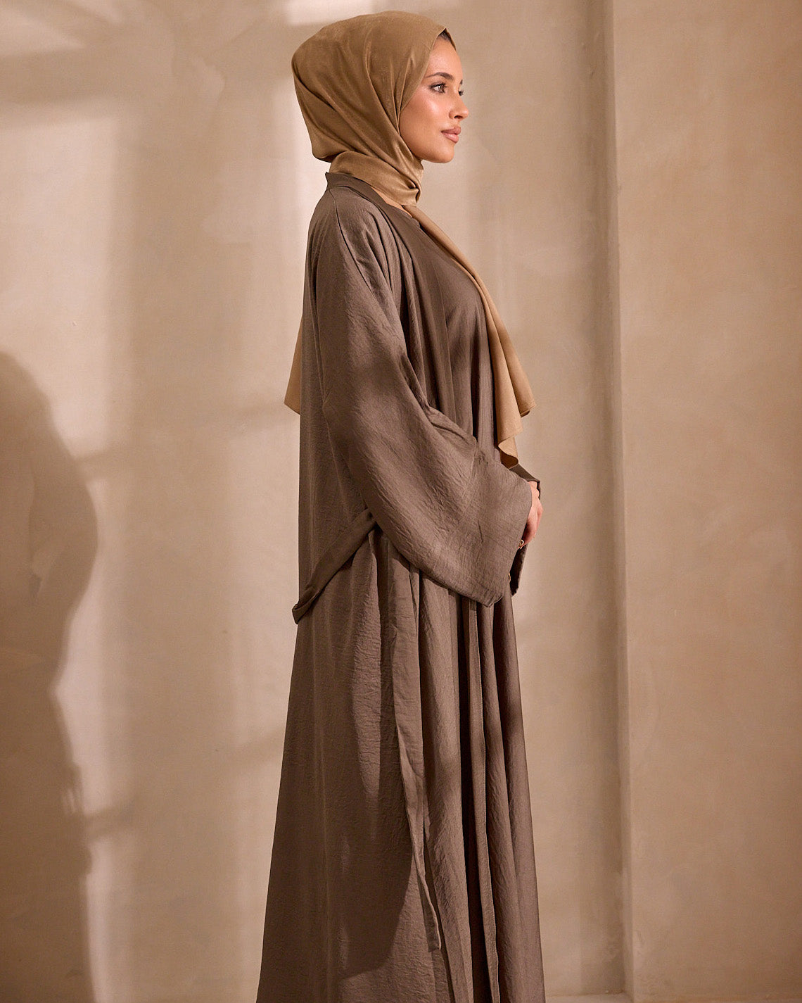 Woman wearing a long brown dress and beige hijab against a beige wall.