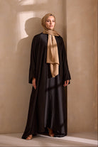 Woman wearing a black abaya and beige hijab against a beige wall.