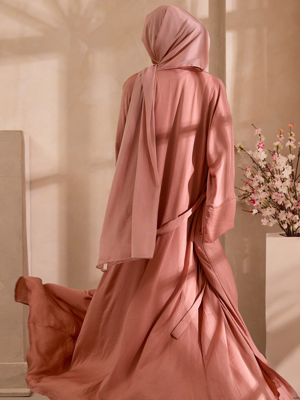 Person wearing a long, flowing pink dress with a matching headscarf in a softly lit room.