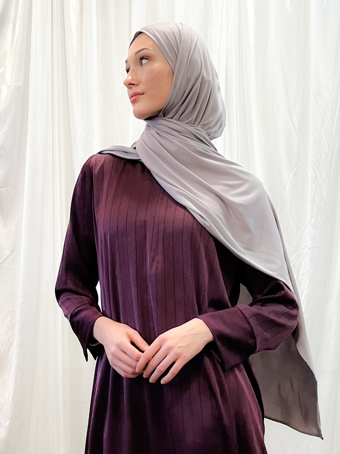 A woman wearing a purple dress and a gray headscarf, standing in front of a white curtain.