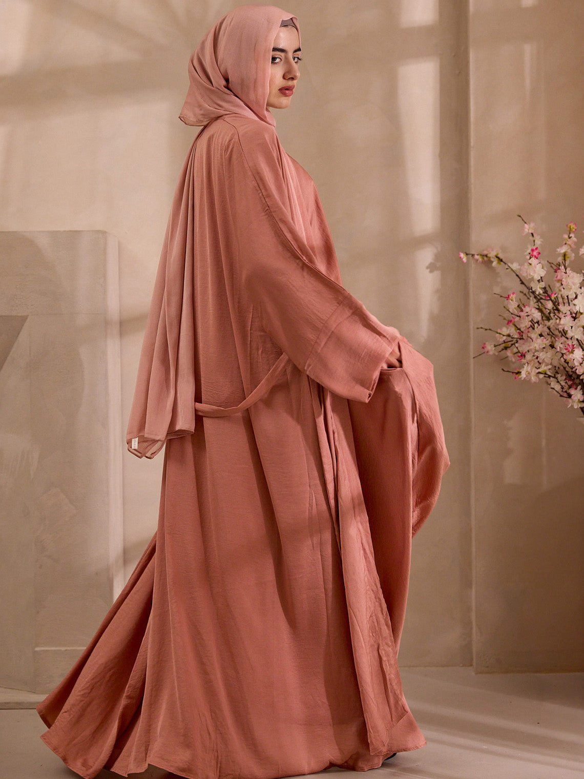 Woman wearing a long, flowing pink dress with a matching hijab in a softly lit room. Abaya Jacket
