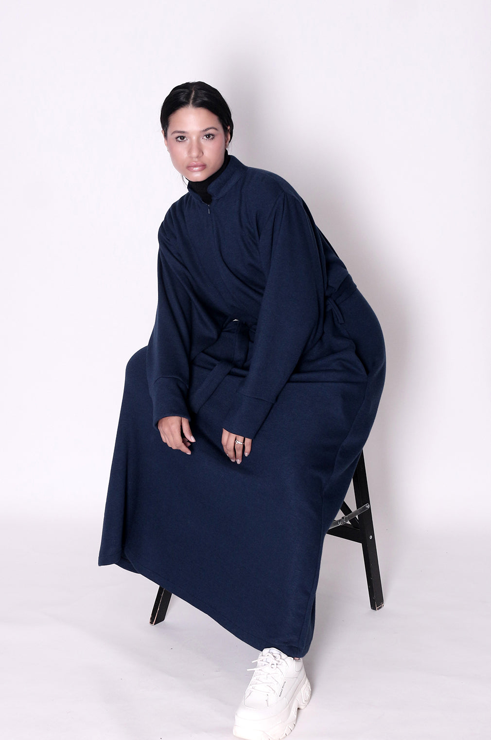 Person wearing a long navy blue garment sitting on a chair against a white background