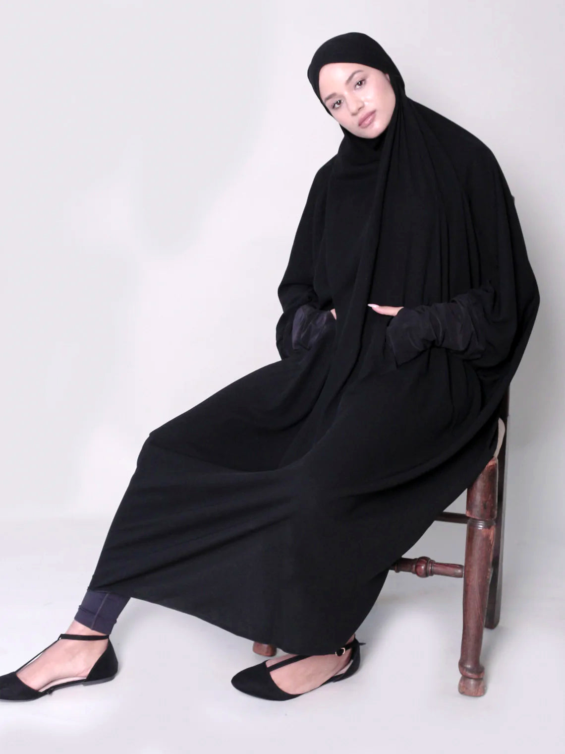 A woman sitting on a chair wearing a black jilbab with thumb holes and a zip at the front.