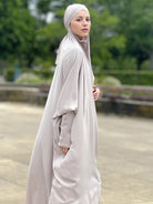 A woman wearing a beige one piece prayer jilbab with a headscarf, standing outdoors. one piece jilbab abaya