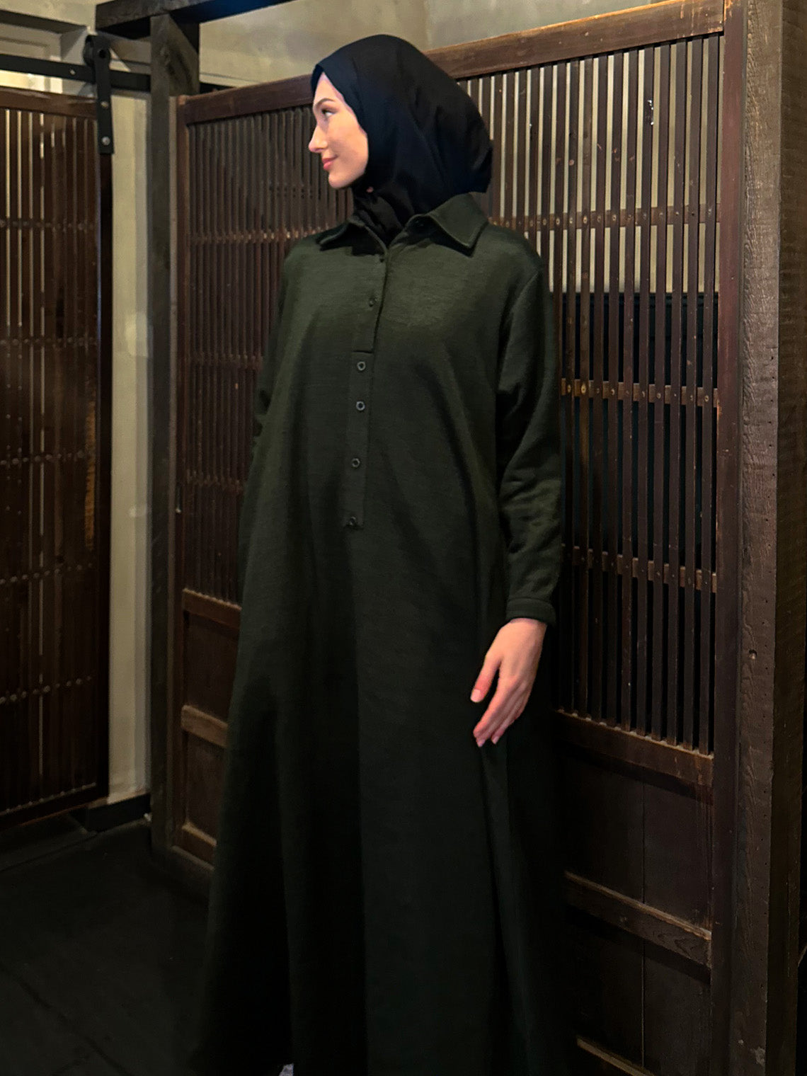 A person standing in front of wooden sliding doors, wearing a khaki knit dress with long sleeves and a round neckline.