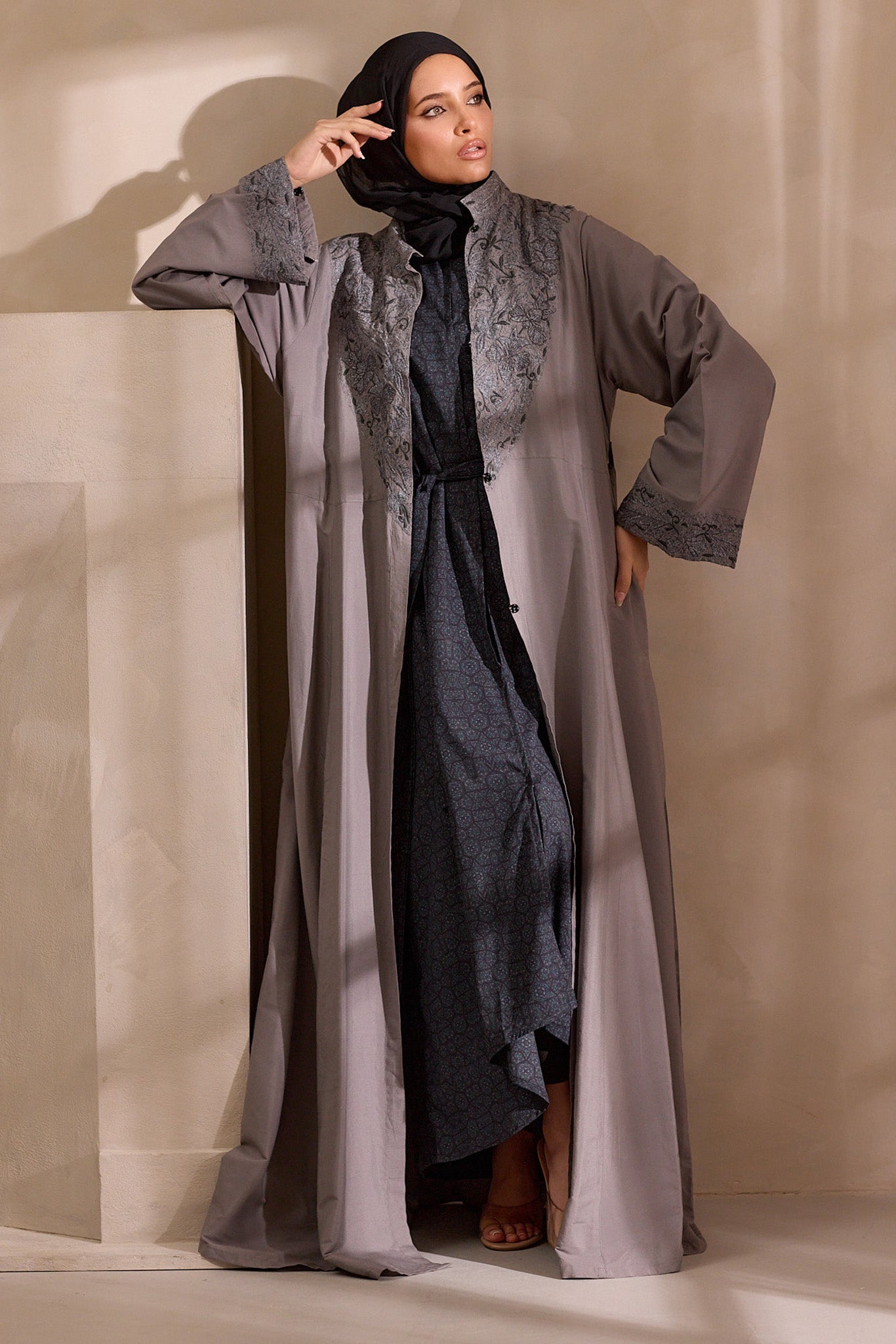 Woman wearing a long, light gray dress with a black hijab against a neutral background open abaya