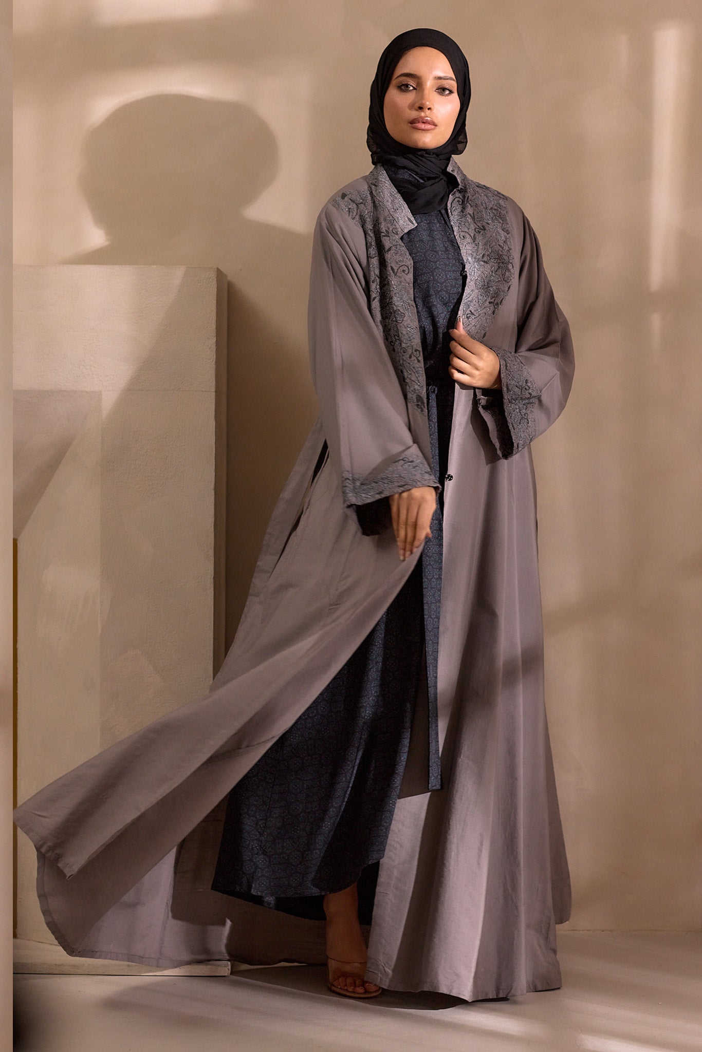 Woman wearing a long, light gray abaya over a dark outfit against a neutral background