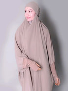Woman wearing a beige hijab and dress against a plain background. one piece jilbab abaya
