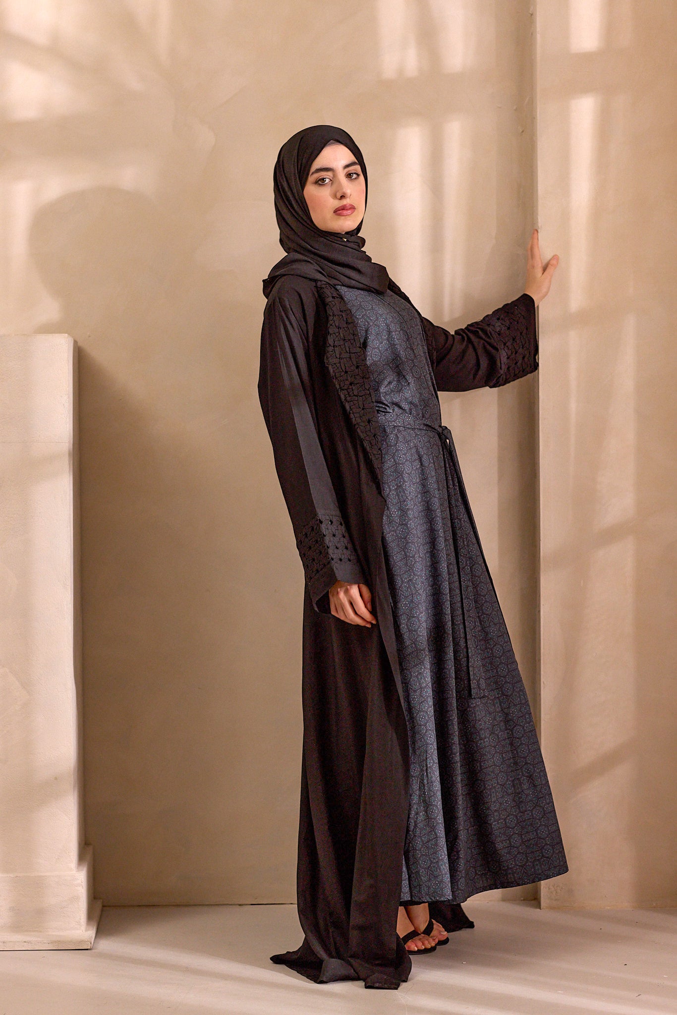 Woman wearing a black and gray abaya against a beige wall.