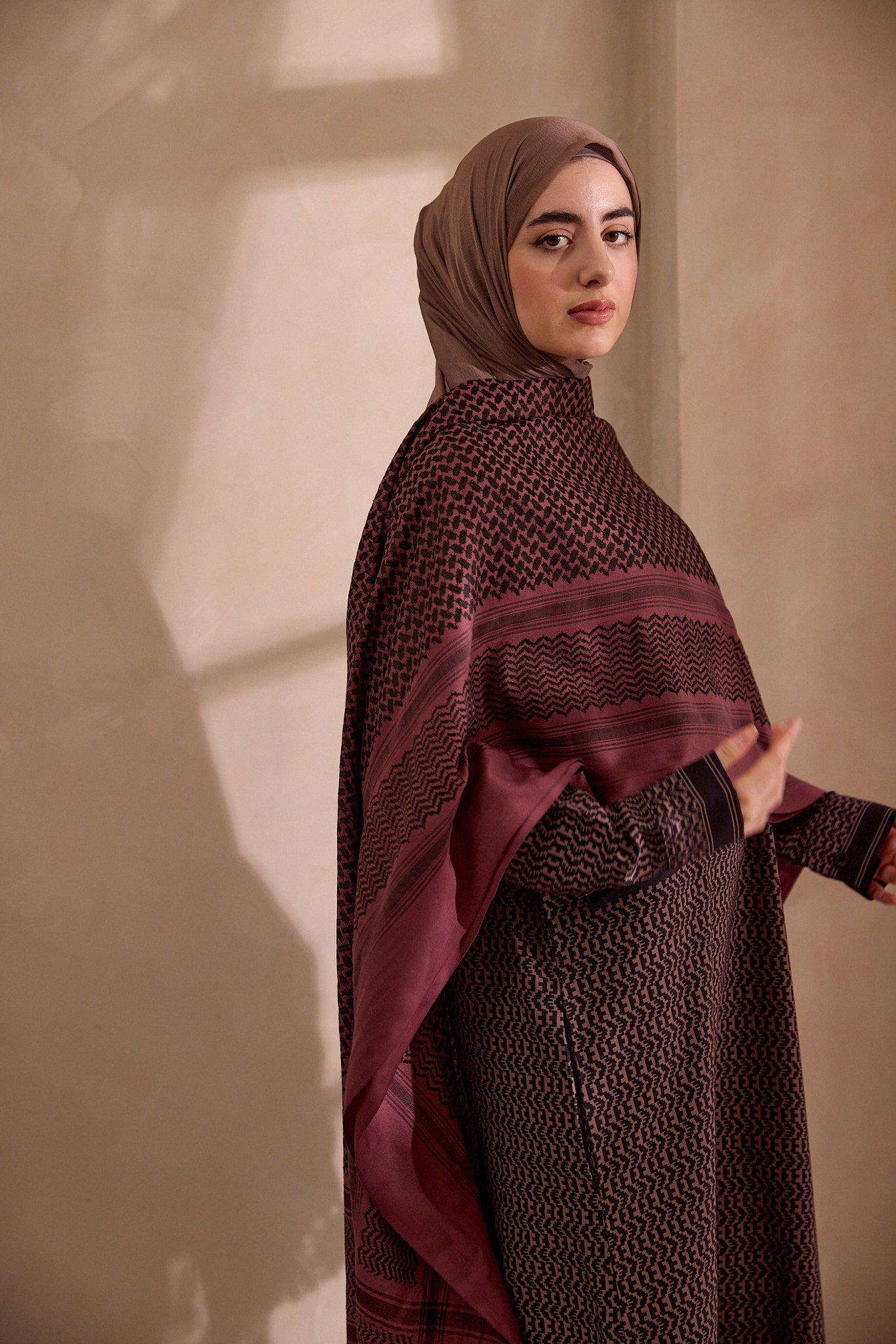 Woman wearing a dark brown hijab and matching outfit against a beige wall.
